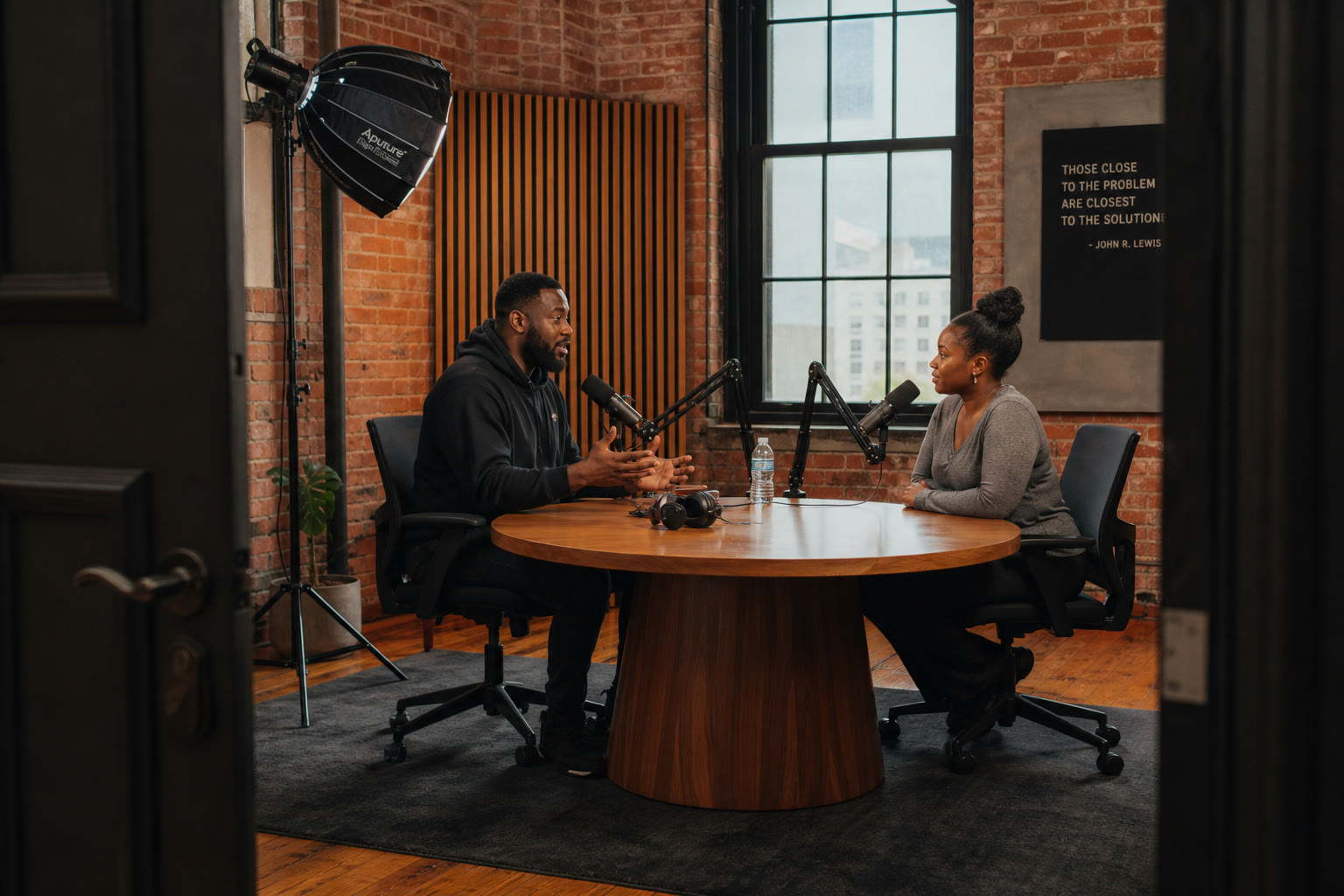 Two people record a conversation at a round table with boom microphones in a brick-walled studio with acoustic wood slats.
